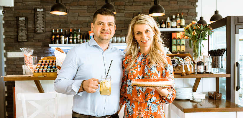 Smiling couple standing together inside a café, holding an iced drink and a tart